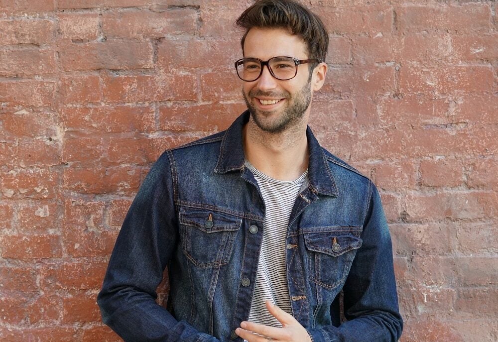 A man wearing glasses and a denim jacket, smiling in front of a brick wall - San Dimas Optometry A man wearing glasses and a denim jacket, smiling in front of a brick wall - San Dimas Optometry