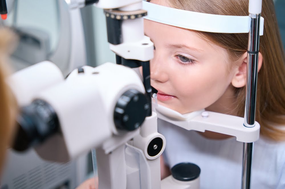 A young girl undergoing an eye examination with her chin placed on the support of a slit lamp machine at an optometrist's office - pediatric eye exam A young girl undergoing an eye examination with her chin placed on the support of a slit lamp machine at an optometrist's office - pediatric eye exam