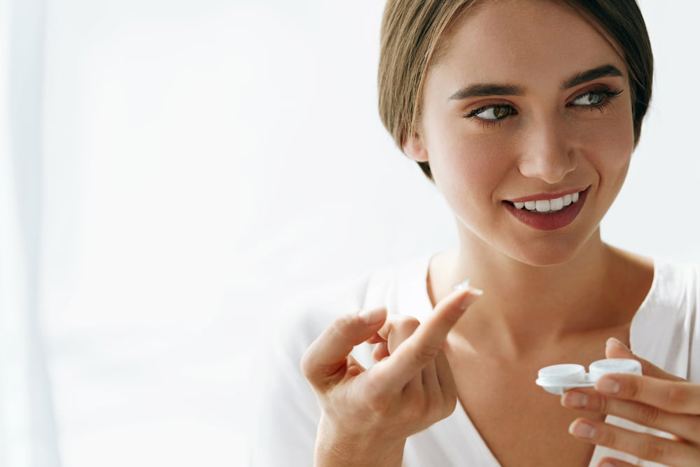 A woman smiling while holding a contact lens case and preparing to place a lens on her finger - contact lens exam A woman smiling while holding a contact lens case and preparing to place a lens on her finger - contact lens exam