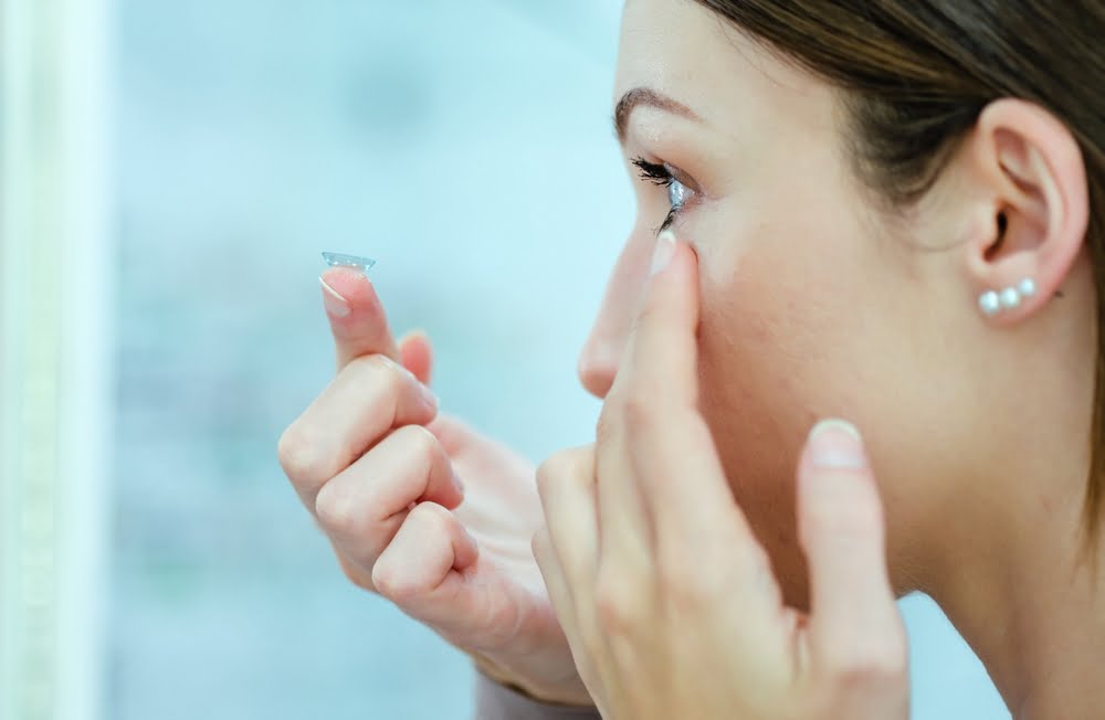 A woman with pearl earrings carefully placing a contact lens in her eye while focusing intently - contact lens exam A woman with pearl earrings carefully placing a contact lens in her eye while focusing intently - contact lens exam