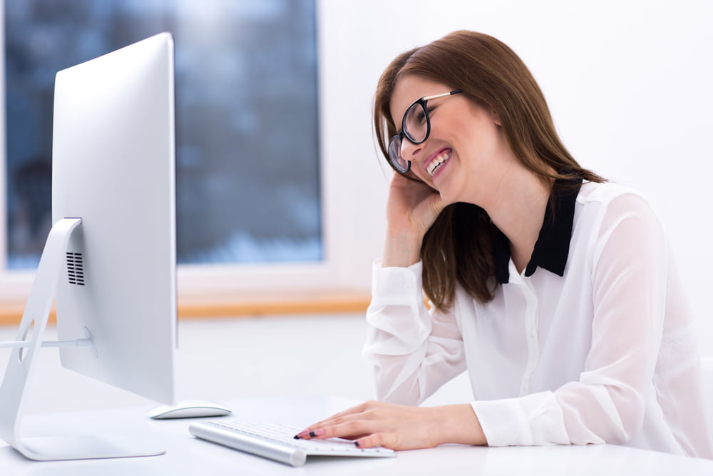 A woman wearing glasses and a white blouse smiles while working at a desktop computer in a bright office - Computer Vision Syndrome A woman wearing glasses and a white blouse smiles while working at a desktop computer in a bright office - Computer Vision Syndrome