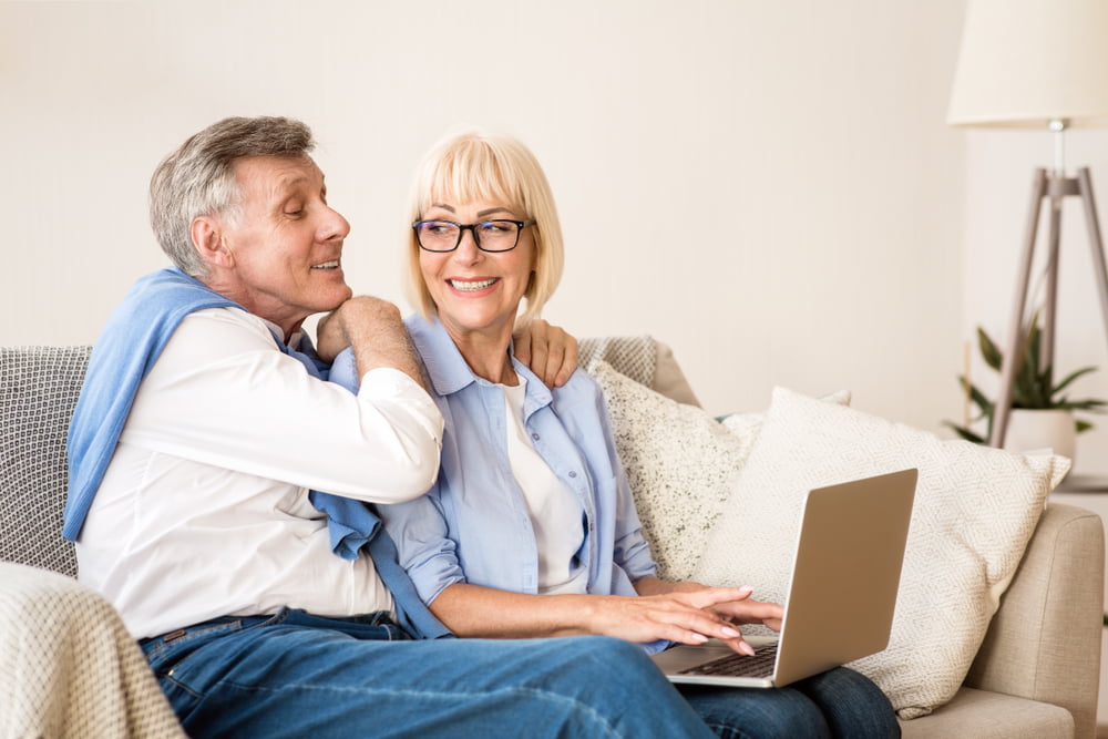 A smiling senior couple sits on a couch together, with the woman using a laptop and the man leaning affectionately towards her - Eye & Vision Exams A smiling senior couple sits on a couch together, with the woman using a laptop and the man leaning affectionately towards her - Eye & Vision Exams