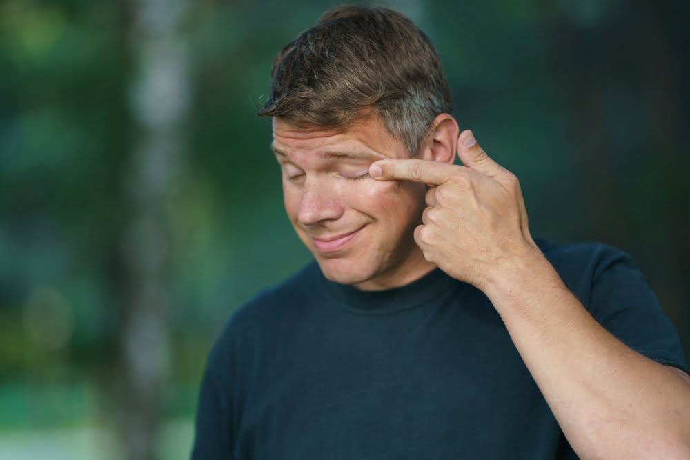 A man outdoors wearing a dark shirt, pointing to his closed eye with his index finger, with a playful or lighthearted expression, against a blurred green background - Eye Allergy Treatment A man outdoors wearing a dark shirt, pointing to his closed eye with his index finger, with a playful or lighthearted expression, against a blurred green background - Eye Allergy Treatment