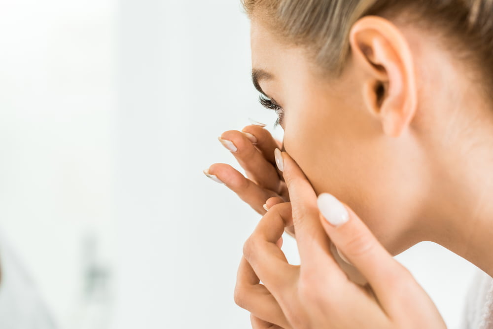 A woman with neatly manicured nails carefully inserting a contact lens into her eye while looking in a mirror - contact lens exam A woman with neatly manicured nails carefully inserting a contact lens into her eye while looking in a mirror - contact lens exam