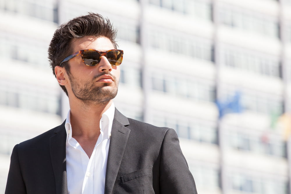 A man in a dark suit and white shirt wears tortoiseshell sunglasses, exuding confidence while standing in front of a modern building - Sunglasses A man in a dark suit and white shirt wears tortoiseshell sunglasses, exuding confidence while standing in front of a modern building - Sunglasses