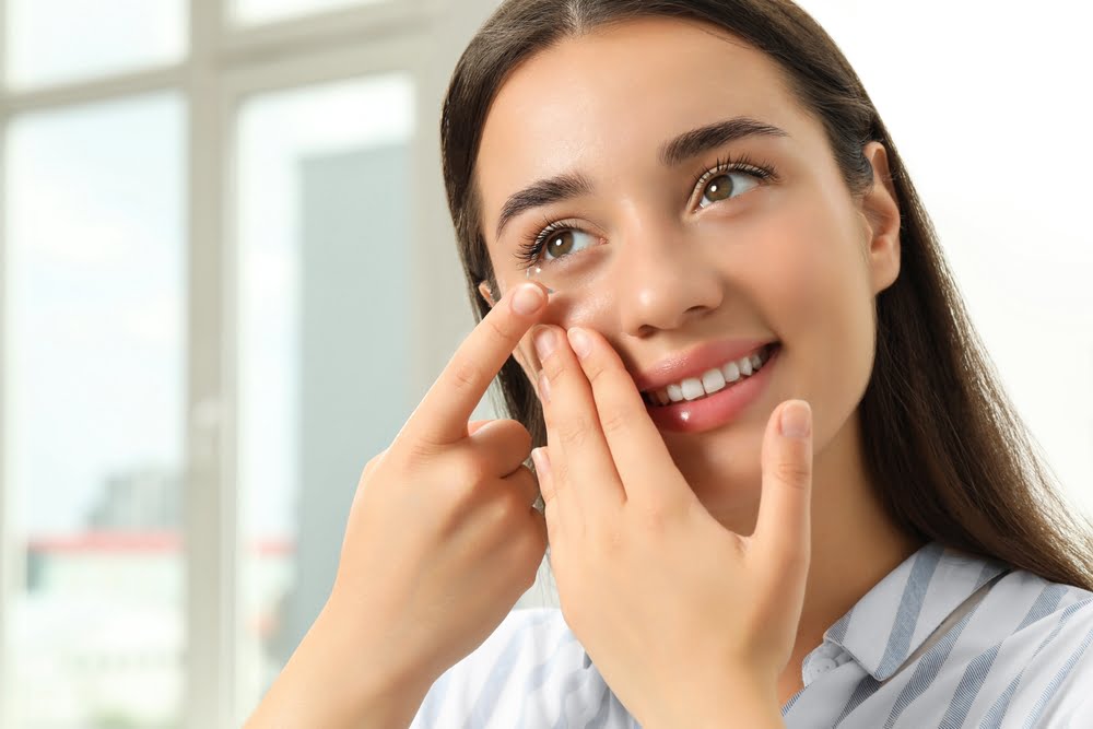 A smiling woman in a striped shirt carefully inserting a contact lens into her eye in a bright room - hard to fit contacts A smiling woman in a striped shirt carefully inserting a contact lens into her eye in a bright room - hard to fit contacts