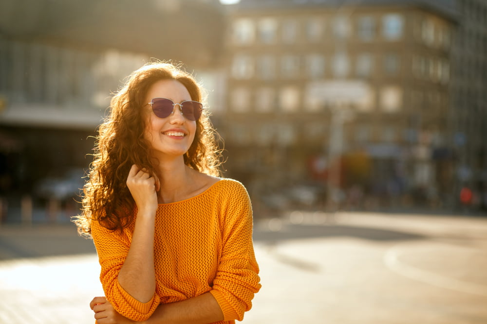 A woman with curly hair, wearing sunglasses and a vibrant yellow sweater, smiles warmly while standing outdoors in sunlight with a blurred urban backdrop - Sunglasses A woman with curly hair, wearing sunglasses and a vibrant yellow sweater, smiles warmly while standing outdoors in sunlight with a blurred urban backdrop - Sunglasses