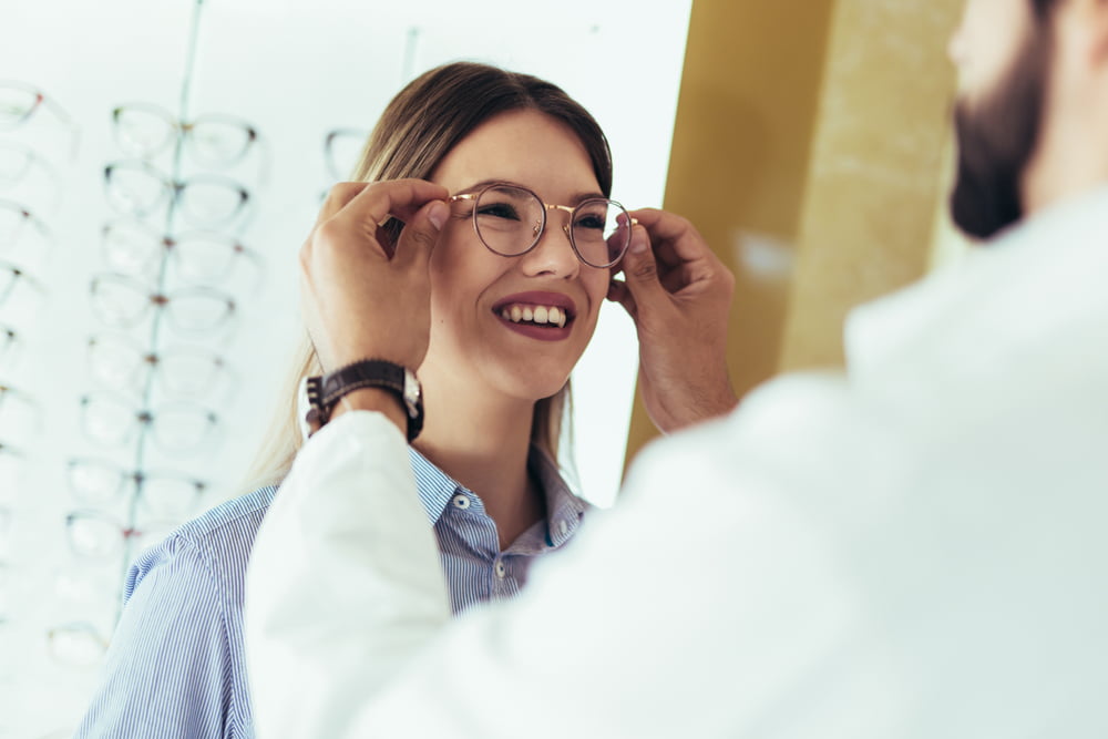 A smiling woman tries on glasses as an optician adjusts the frames - Myopia Control A smiling woman tries on glasses as an optician adjusts the frames - Myopia Control