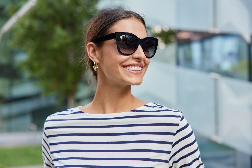 A smiling woman wearing oversized black sunglasses, gold earrings, and a striped shirt stands outdoors against a modern glass building with greenery in the background - Sunglasses A smiling woman wearing oversized black sunglasses, gold earrings, and a striped shirt stands outdoors against a modern glass building with greenery in the background - Sunglasses