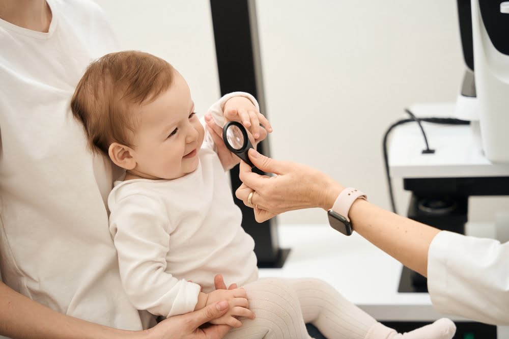Pediatric Eye Exam in San Dimas, CAaA smiling baby sits on an adult's lap while a healthcare professional shows them a handheld lens during an eye check-up - pediatric eye exam A smiling baby sits on an adult's lap while a healthcare professional shows them a handheld lens during an eye check-up - pediatric eye exam