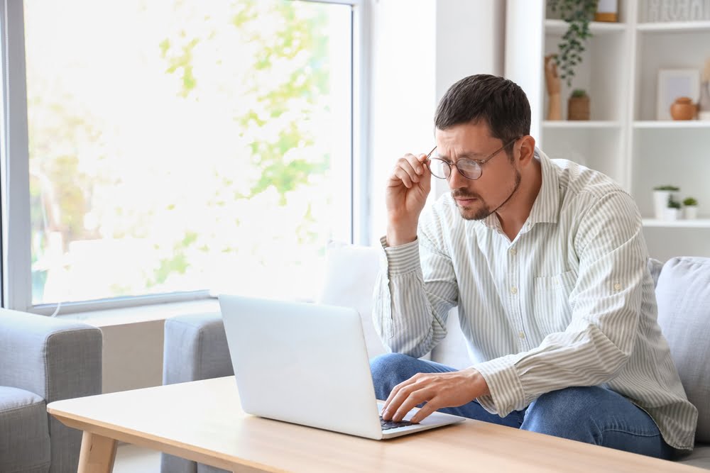 A man sitting on a couch in a well-lit room, adjusting his glasses while focused on a laptop placed on a wooden coffee table - Computer Vision Syndrome A man sitting on a couch in a well-lit room, adjusting his glasses while focused on a laptop placed on a wooden coffee table - Computer Vision Syndrome