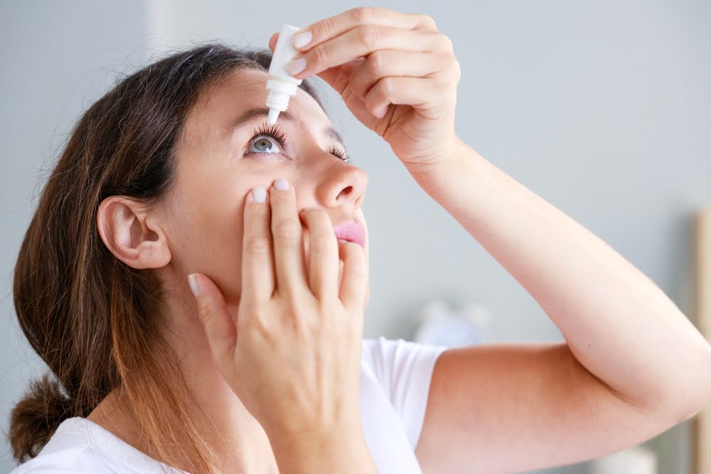 A woman in a white shirt tilting her head back and holding an eye drop bottle above her open eye, using her other hand to gently pull down her lower eyelid, with a soft indoor background - Eye Allergy Treatment A woman in a white shirt tilting her head back and holding an eye drop bottle above her open eye, using her other hand to gently pull down her lower eyelid, with a soft indoor background - Eye Allergy Treatment