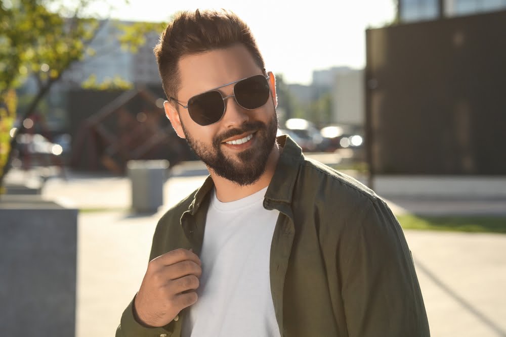 A bearded man wearing sunglasses, a white t-shirt, and a green jacket smiles confidently outdoors with a blurred urban setting and sunlight in the background - Sunglasses A bearded man wearing sunglasses, a white t-shirt, and a green jacket smiles confidently outdoors with a blurred urban setting and sunlight in the background - Sunglasses