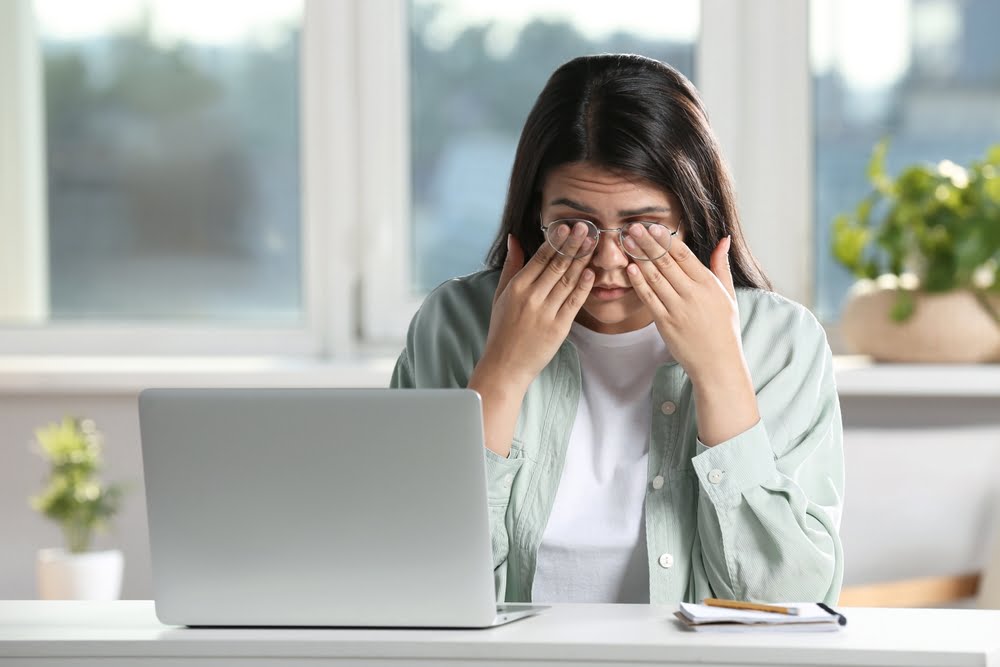 A woman sitting at a desk in a bright room, rubbing her eyes while working on a laptop with a notebook and pen nearby - Computer Vision Syndrome A woman sitting at a desk in a bright room, rubbing her eyes while working on a laptop with a notebook and pen nearby - Computer Vision Syndrome