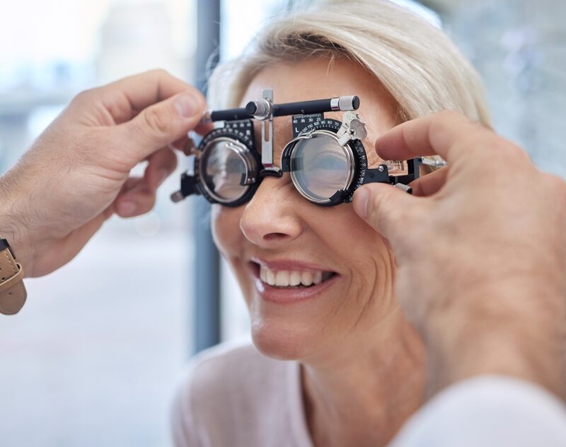 A smiling woman undergoing an eye exam as an optometrist adjusts the lenses on a trial frame - Eye & Vision Exams
