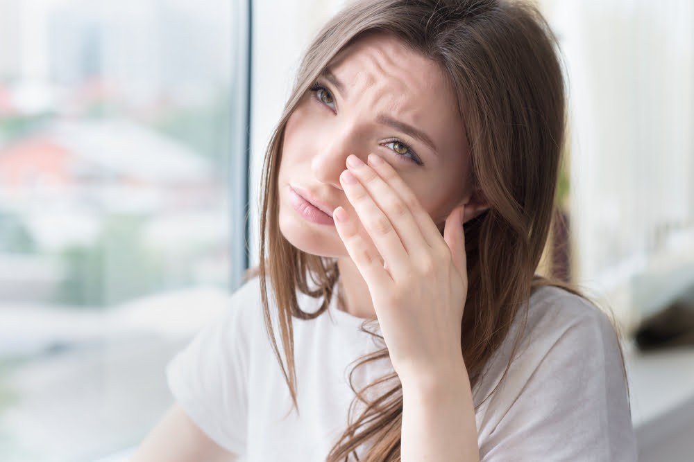 A woman rubs her eye, appearing uncomfortable as she looks out of a window - Eye allergy treatment 