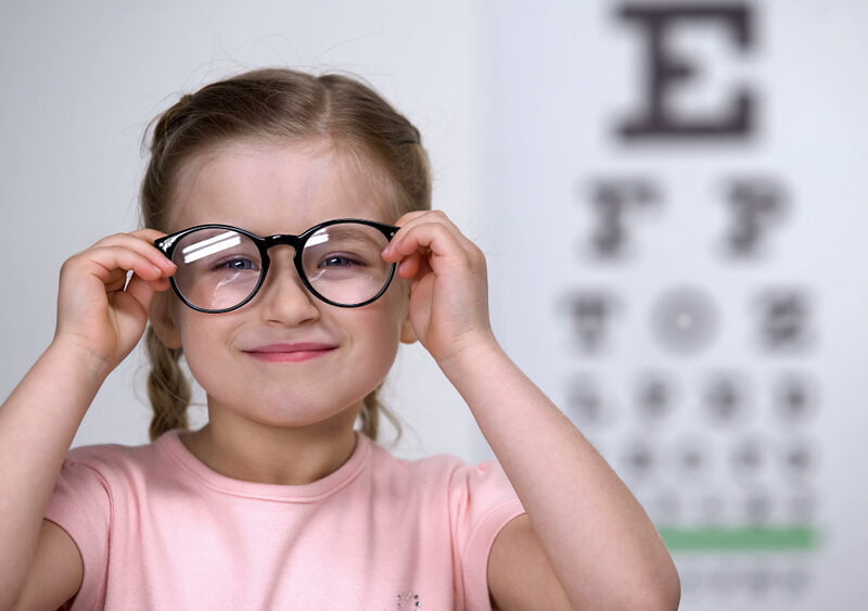 A young girl wearing oversized glasses smiles in front of an eye chart at an optometry clinic - Pediatric eye exams