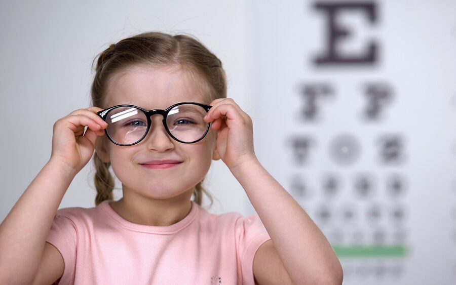 A young girl wearing oversized glasses smiles in front of an eye chart at an optometry clinic - Pediatric eye exams