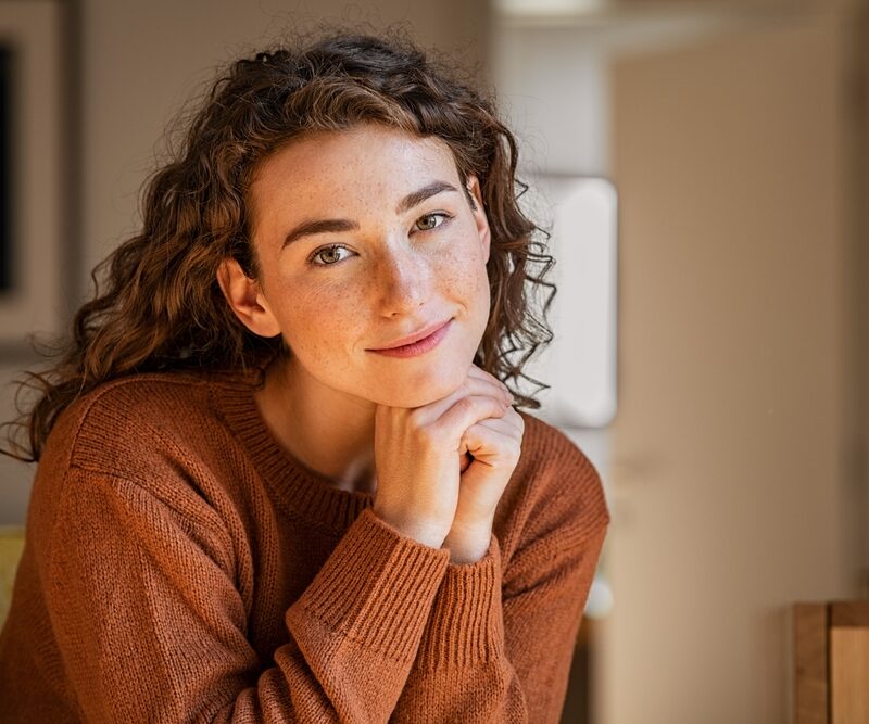 A woman with curly hair wearing a brown sweater smiles gently while resting her chin on her hand - Specialty contact lens