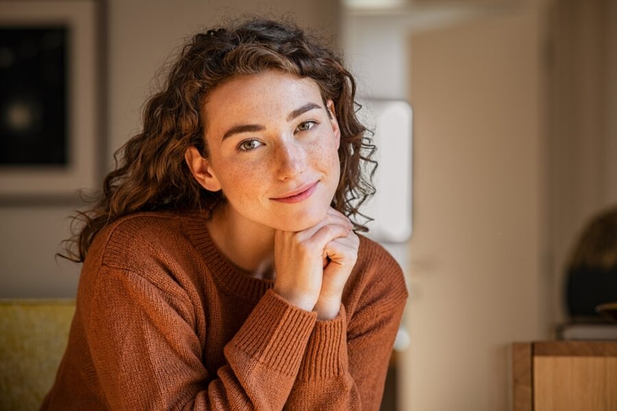 A woman with curly hair wearing a brown sweater smiles gently while resting her chin on her hand - Specialty contact lens